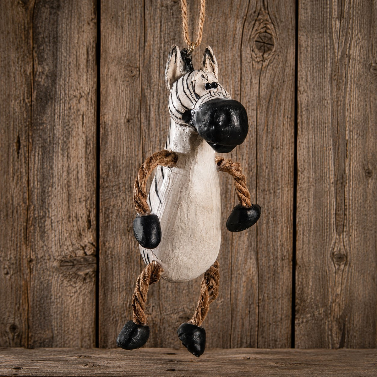 A hanging zebra plush toy or decoration made from a white stuffed body with black painted stripes, a black snout and hooves, and rope legs, shown against a rustic wooden plank background.