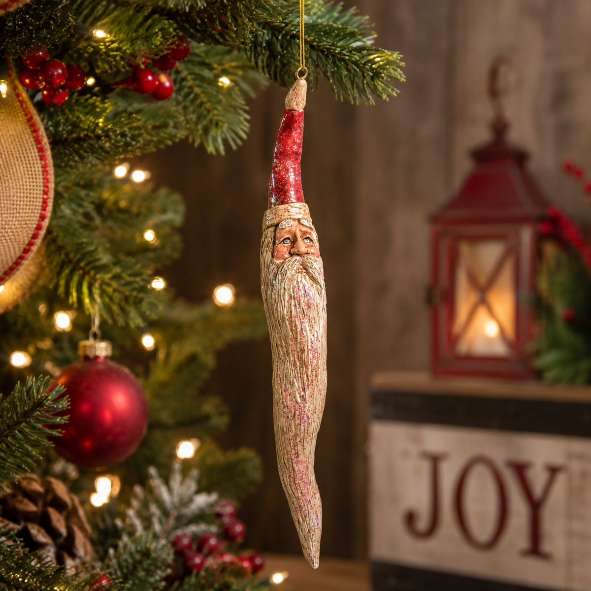 A rustic Santa Claus Christmas ornament shaped like a hanging icicl, suspended from a decorated Christmas tree with warm lights and red ornaments.