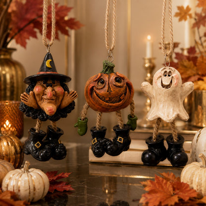 Three Halloween-themed hanging ornaments displayed on a tabletop: a witch figure in a black hat and boots, a carved jack-o'-lantern with green hands, and a white ghost with black boots. The scene is styled with autumn leaves, small pumpkins, candles, and warm background decor.