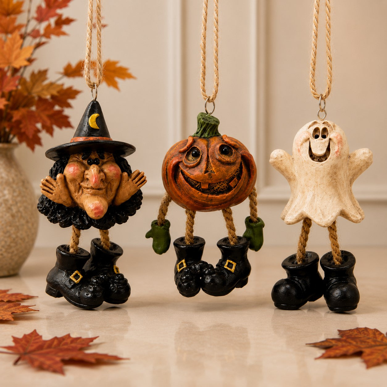 Three Halloween hanging ornaments shaped like a witch, a jack-o'-lantern, and a ghost, each with a rope hanger and black boots, displayed with autumn leaves and a vase in the background.