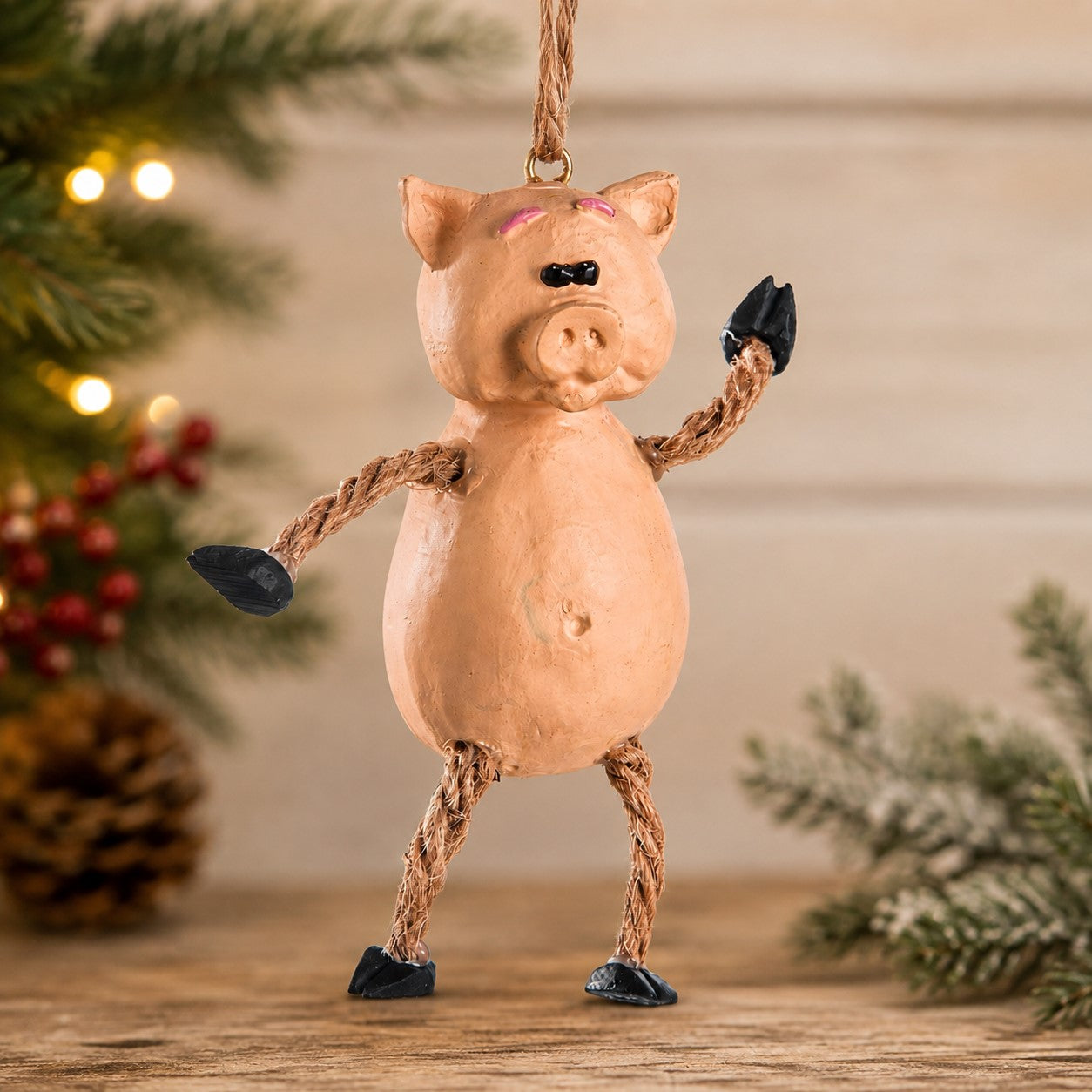 A handmade pig-shaped Christmas ornament with a peach-colored body, rope arms and legs, and black hooves, hanging on a wooden surface with a festive holiday background.