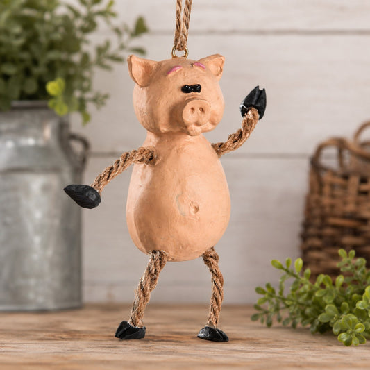 A rustic hanging pig decoration made from a peach-colored wooden or clay body with rope arms and legs and black hooves, displayed on a wooden surface with a blurred farmhouse-style background.