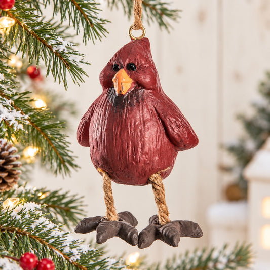 Bert Anderson cardinal ornament with dangling rope legs displayed on a bright Christmas tree with snowy evergreen branches, red berries and warm lights.