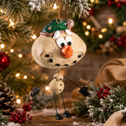 A resin Christmas snowman ornament hanging from a decorated Christmas tree. The ornament features a beige body, black stick-like limbs, a green hat, and a bright orange nose, set against a festive holiday background with warm lights and pine branches.