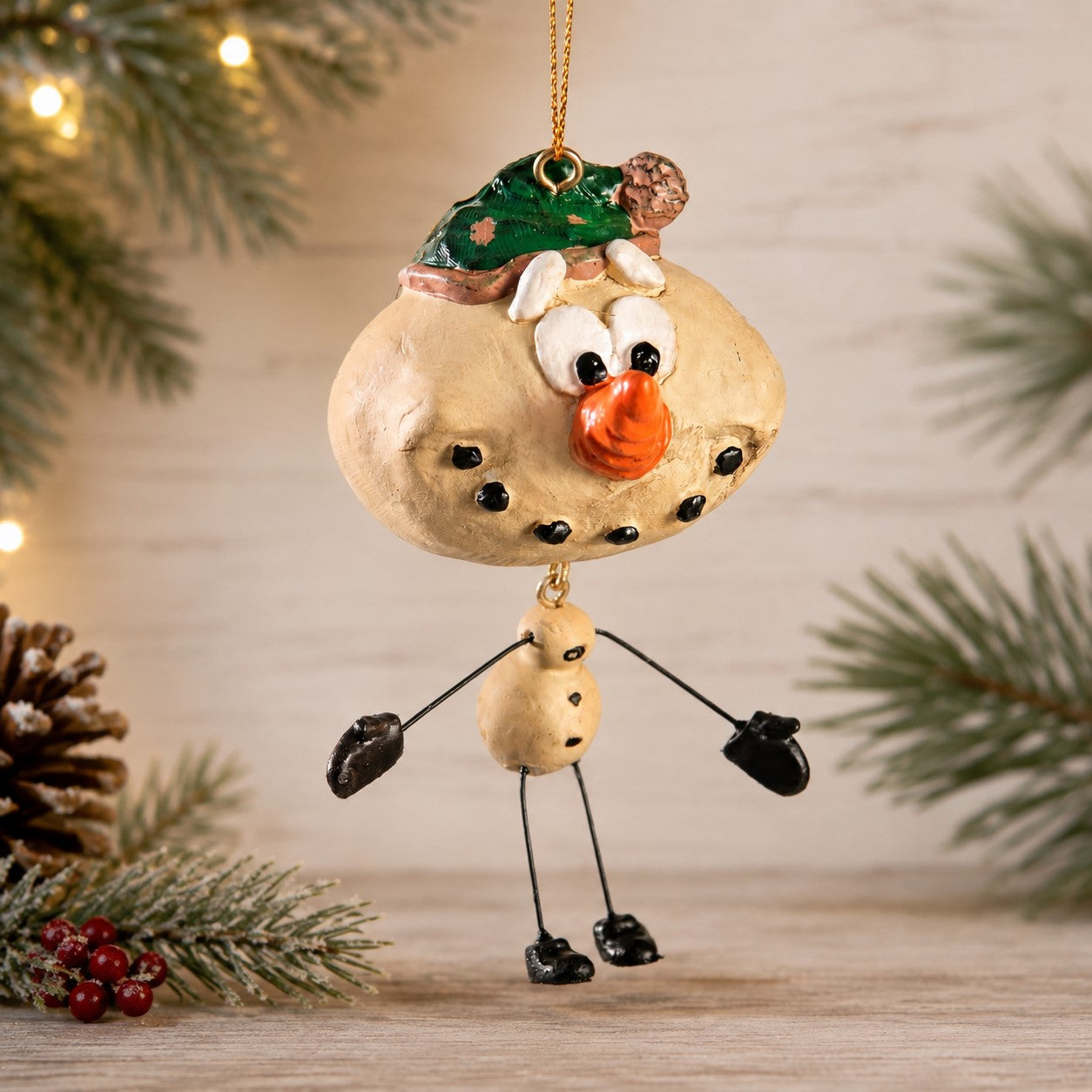 A resin Christmas ornament shaped like a smiling snowman with a green hat, standing on black stick legs with black mittens, photographed against a softly blurred holiday background with pine branches and warm lights.