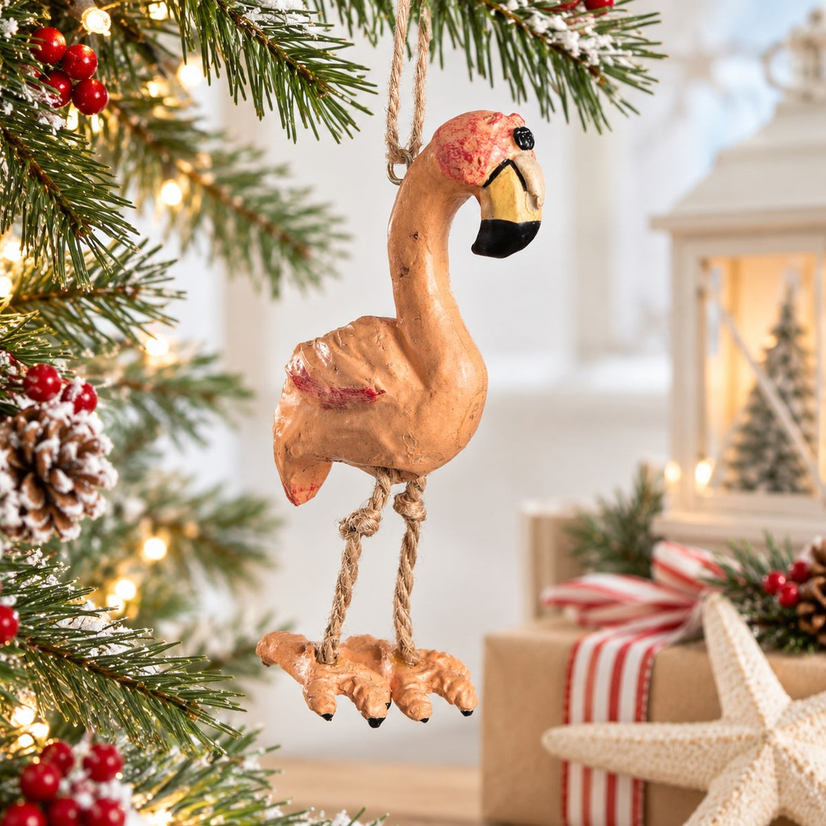 Bert Anderson flamingo ornament with dangling rope legs displayed on a bright Christmas tree with plaid ribbon, red berries and warm holiday lights.