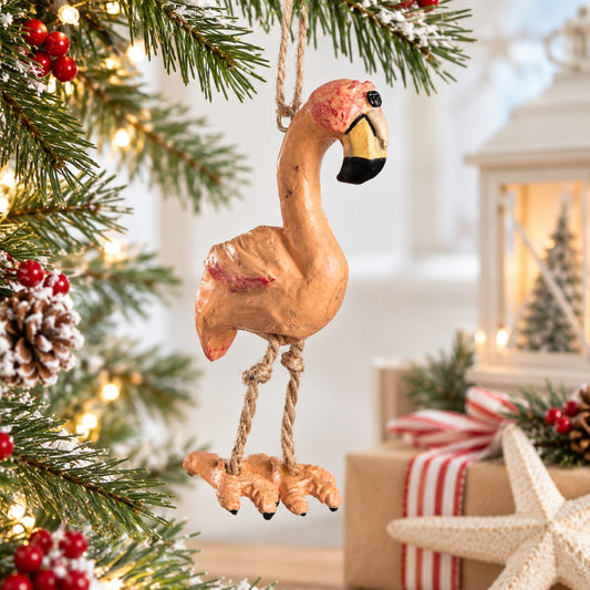 Bert Anderson flamingo ornament with dangling rope legs displayed on a bright Christmas tree with plaid ribbon, red berries and warm holiday lights.