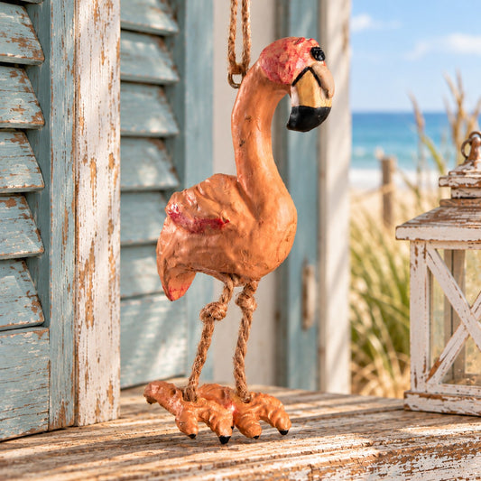 Bert Anderson flamingo ornament with dangling rope legs displayed against a weathered coastal beach hut with blue shutters and an ocean view.