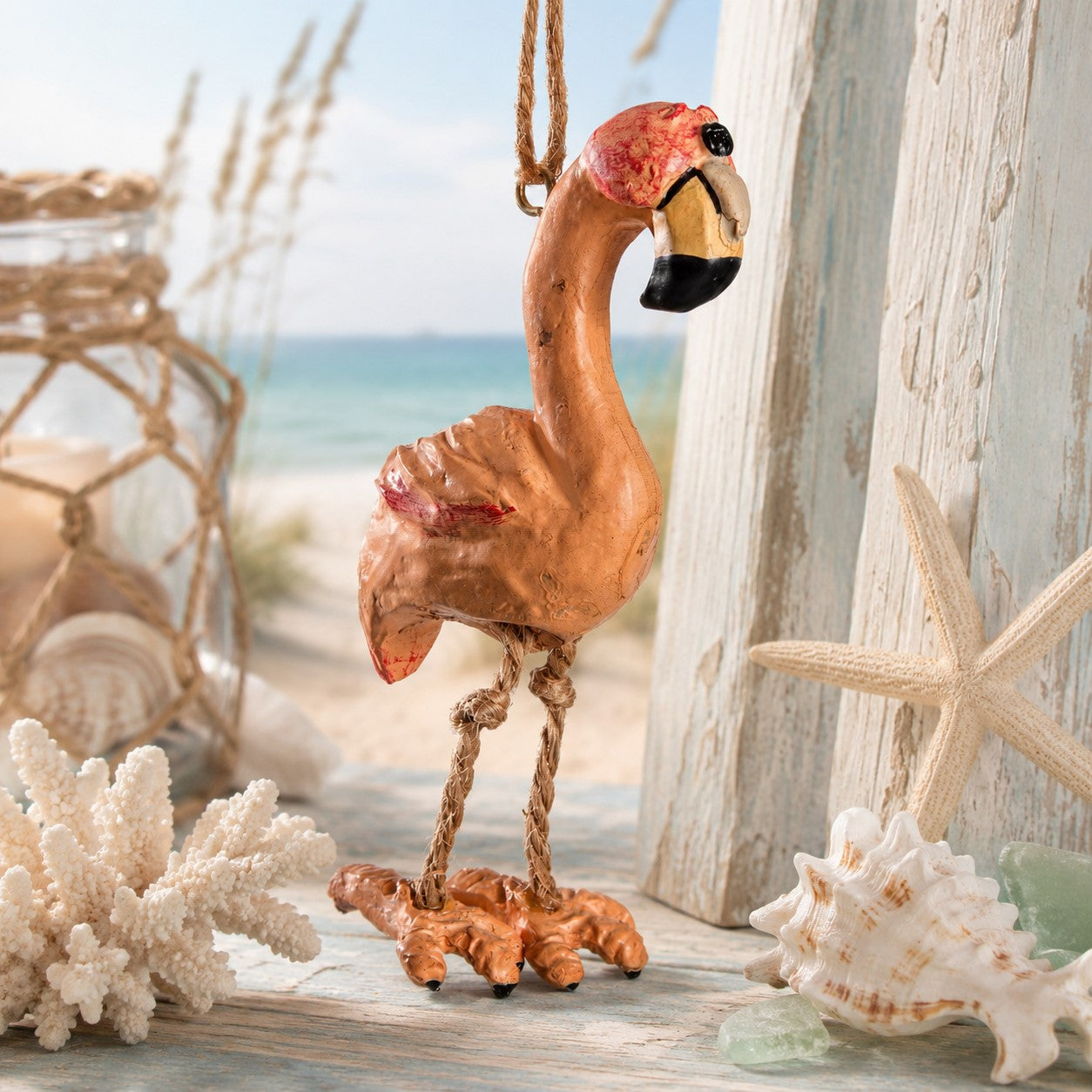 A decorative flamingo ornament hanging from a rope, styled on a rustic wooden surface with seashells, coral, and a beach scene in the background.