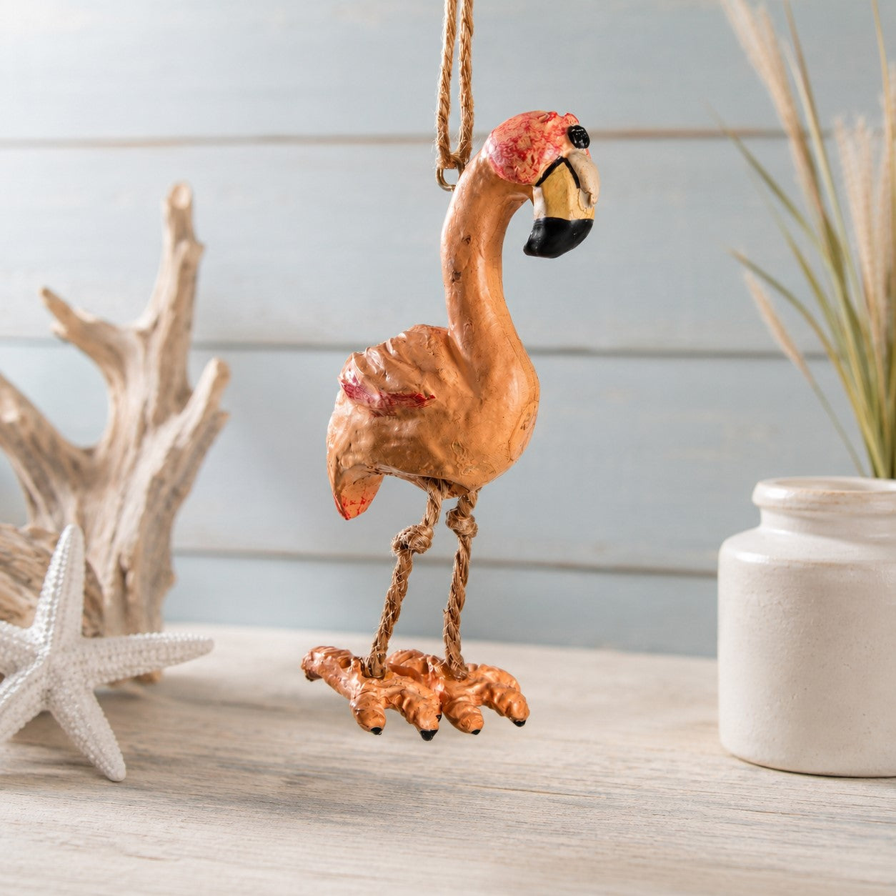 A decorative flamingo hanging ornament in a warm orange-pink tone with red and black accents on its beak and head, suspended by a rope against a light blue wood plank background with driftwood, a starfish, and a white vase nearby.