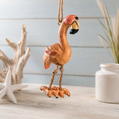 A decorative flamingo hanging ornament in a warm orange-pink tone with red and black accents on its beak and head, suspended by a rope against a light blue wood plank background with driftwood, a starfish, and a white vase nearby.