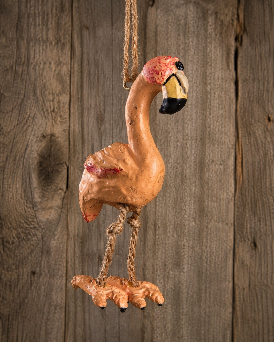 A hand-painted resin flamingo ornament hanging from a rope against a rustic wooden plank background.