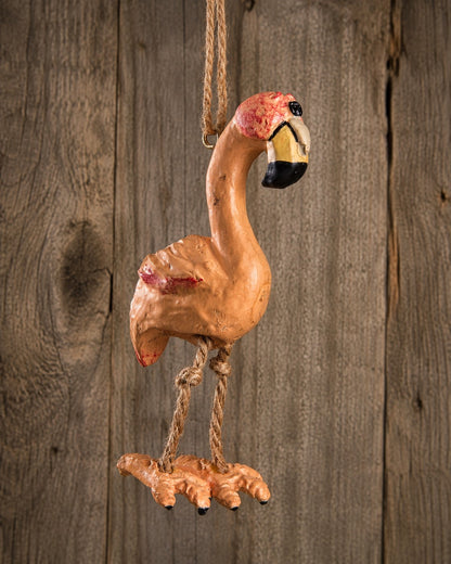 A hand-painted resin flamingo ornament hanging from a rope against a rustic wooden plank background.