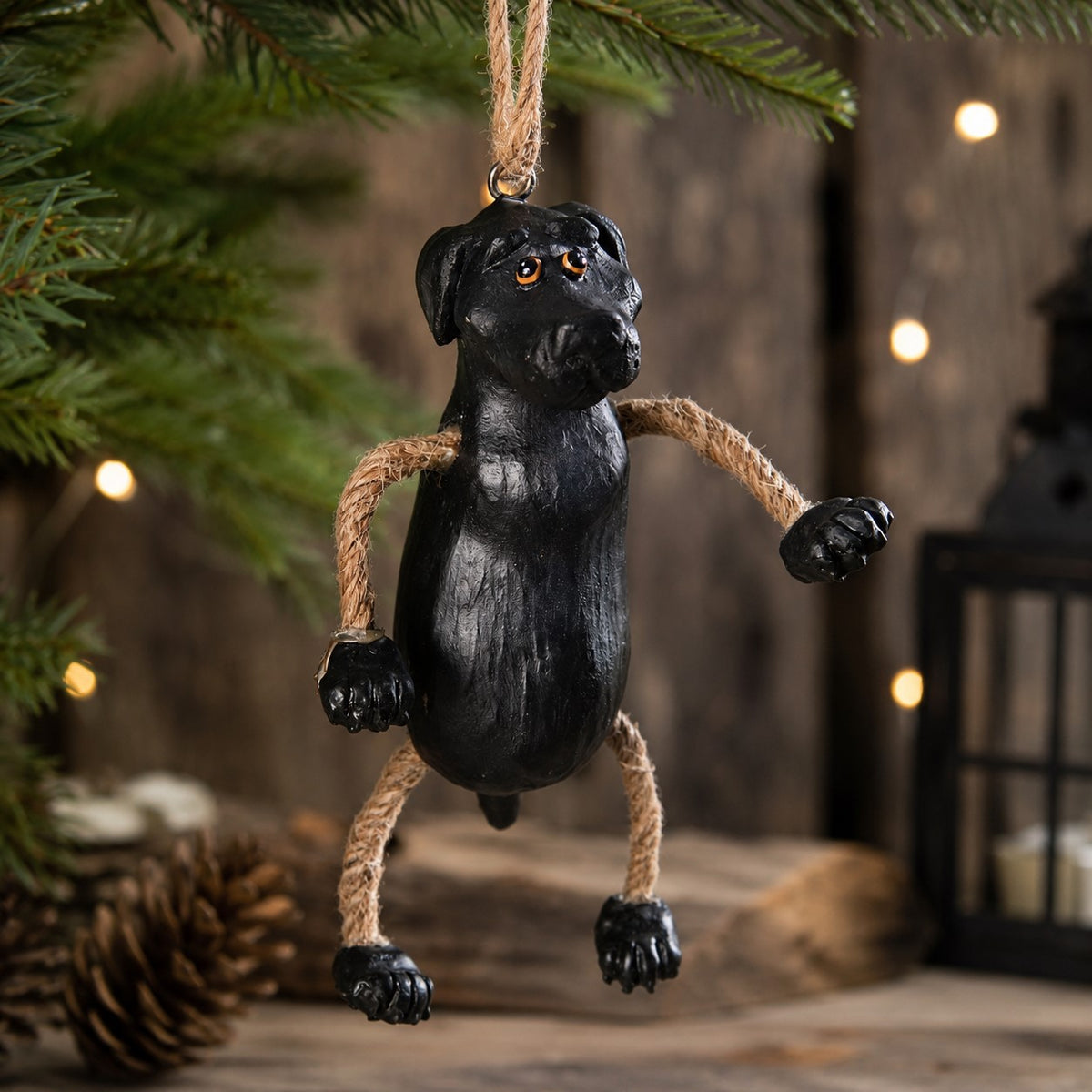 A black dog hanging ornament with rope limbs and a hanging twine loop, displayed on a Christmas tree branch with warm string lights in the background.