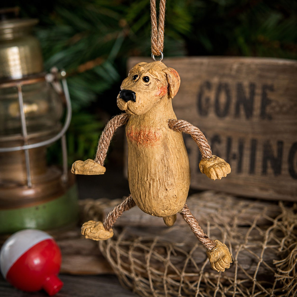 A rustic resin dog hanging ornament with rope limbs and a hanging loop, styled on a wooden surface with a lantern, net, and a blurred sign in the background.