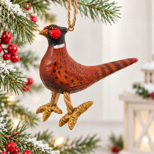Bert Anderson pheasant ornament with dangling feet displayed on a bright Christmas tree with snowy evergreen branches, red berries, pinecones and warm lights.