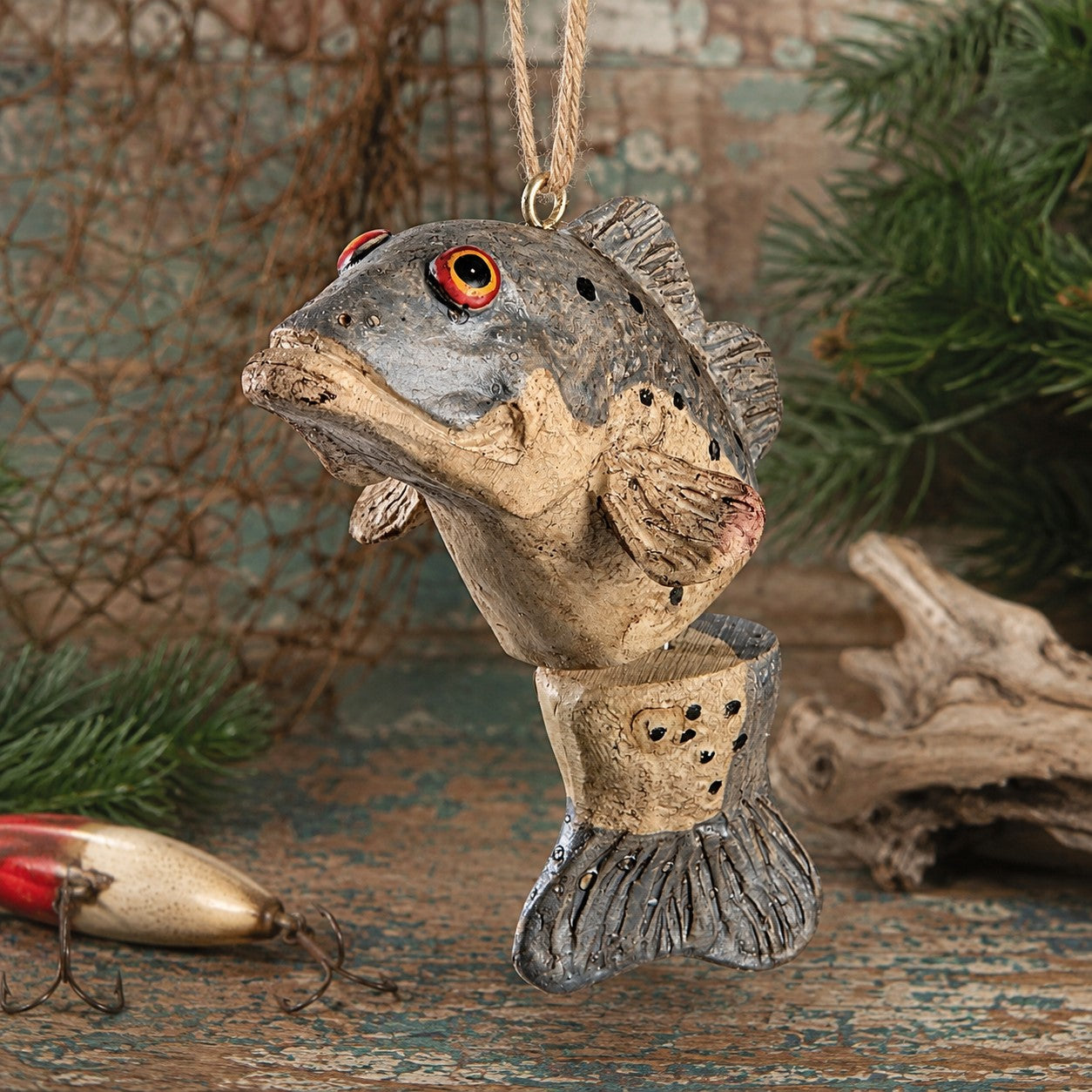 A bass fish hanging ornament with a textured gray and beige body, red eyes, and a natural twine loop at the top, displayed on a rustic wooden surface with driftwood, evergreen branches, and a fishing lure in the background.