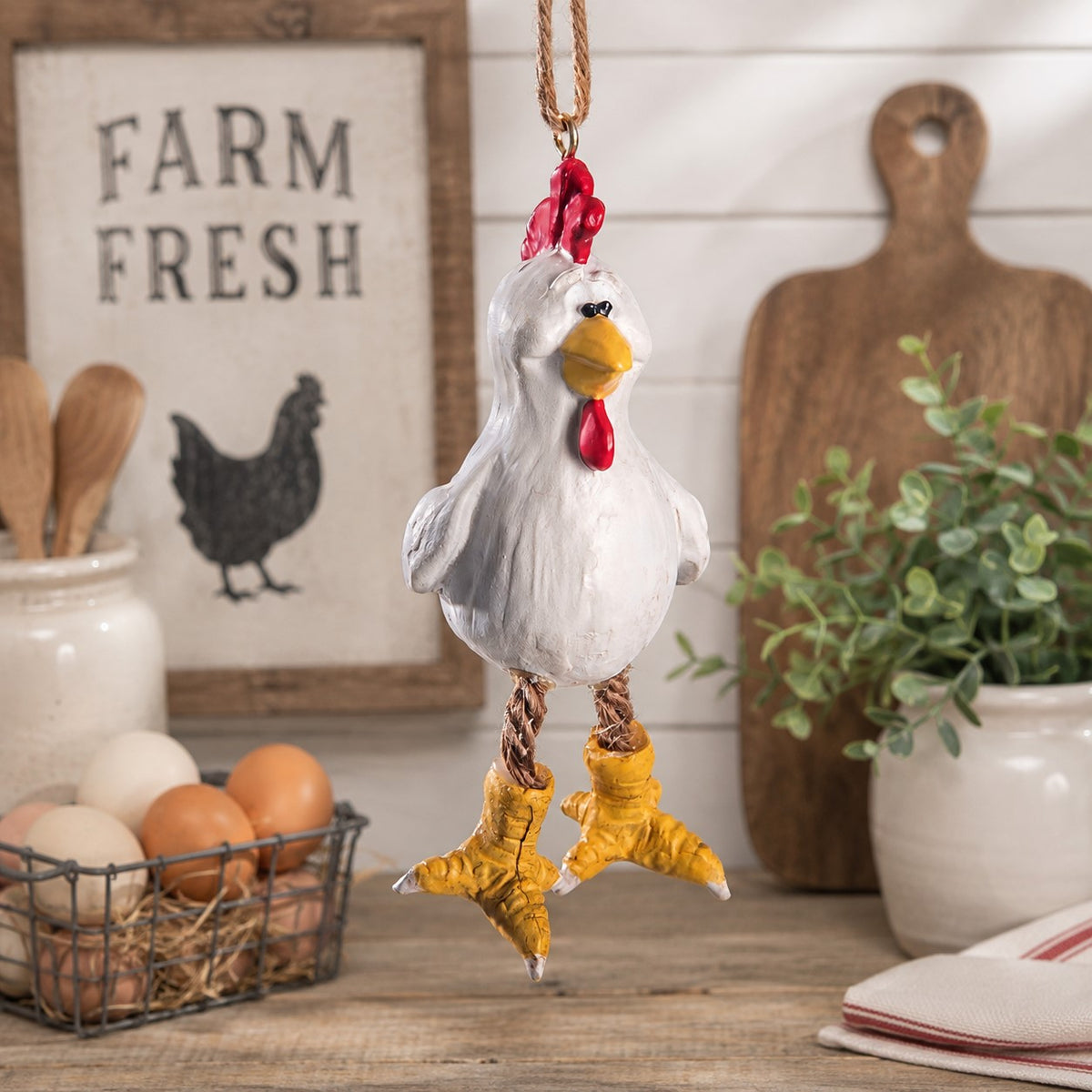 A hanging chicken ornament with a white body, red comb and wattle, yellow beak and feet, and rope-wrapped legs, displayed in a rustic kitchen with farm-themed decor in the background.