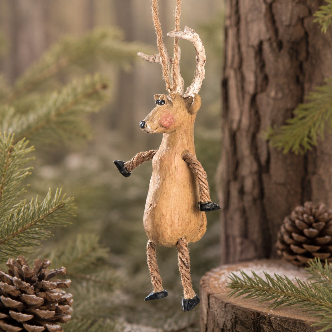 A hanging deer ornament made from resin and rope, with antlers, hanging from a tree stump in a forest setting with pine branches and a pinecone.