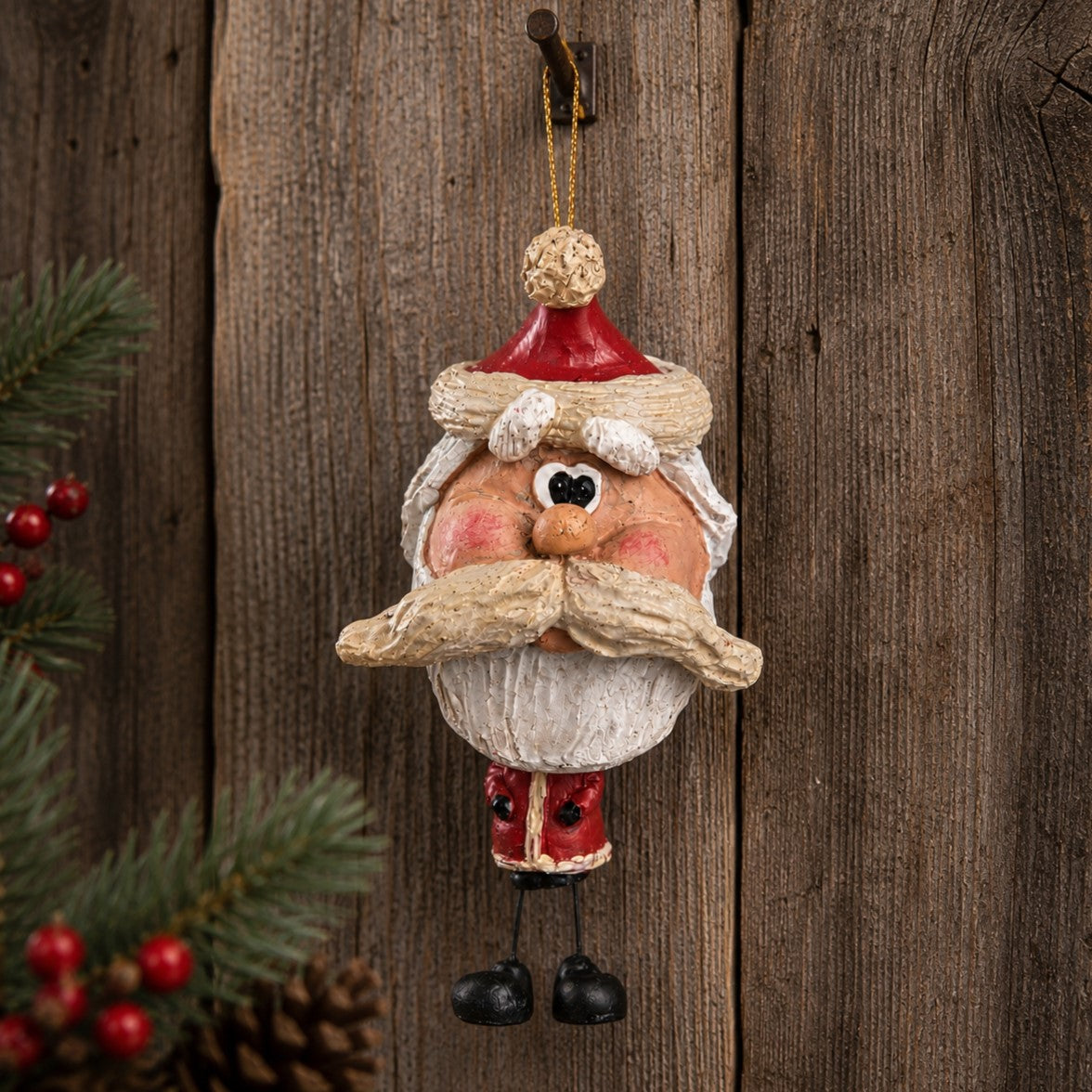 A Santa Claus hanging ornament with a textured beige beard and mustache, red hat and coat, black shoes, and a gold hanging loop, shown against a rustic wooden background with pine branches and red berries at the side.