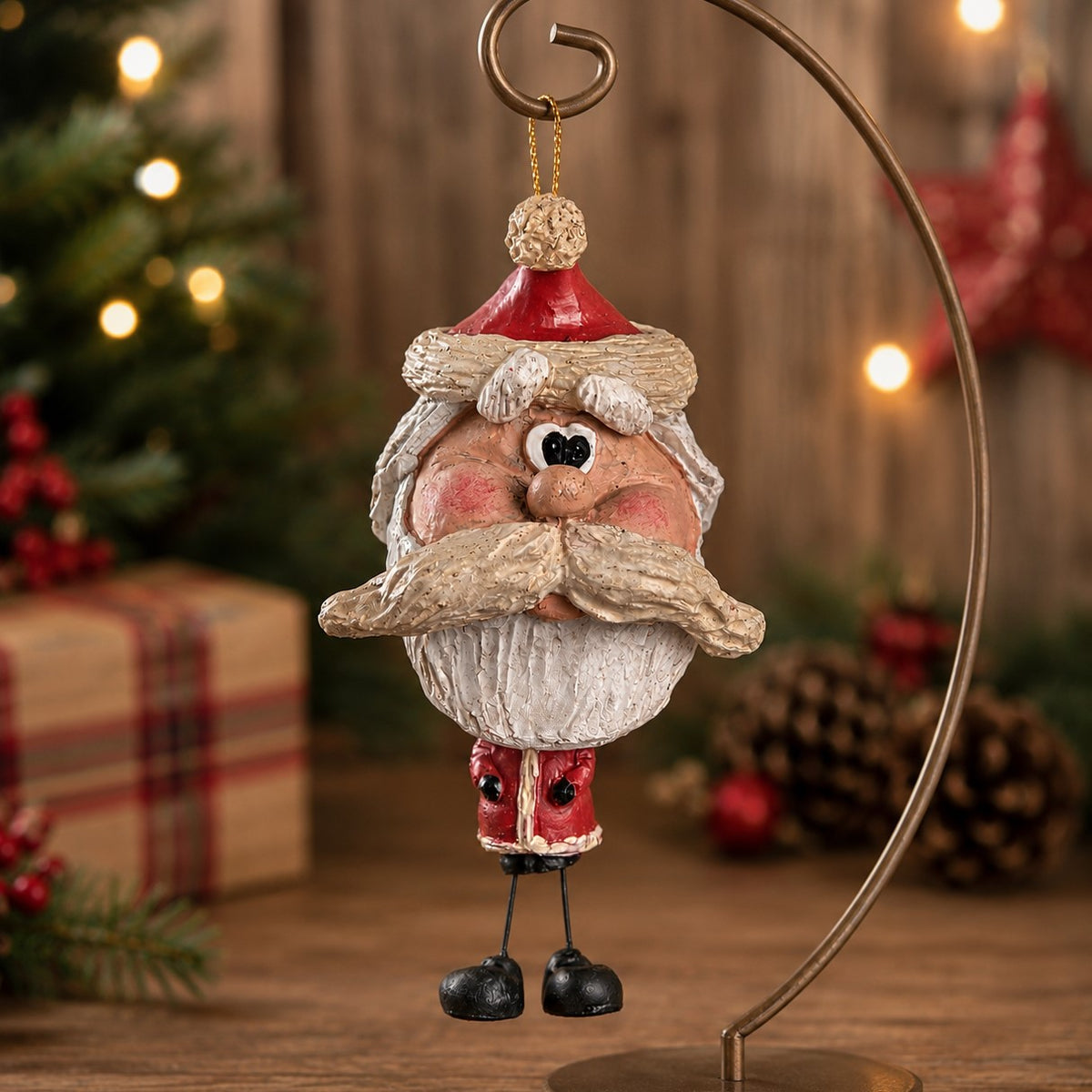A Santa Claus hanging ornament with a rustic, weathered finish, displayed on a wooden surface with a blurred Christmas tree and holiday decorations in the background.
