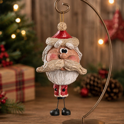 A Santa Claus hanging ornament with a rustic, weathered finish, displayed on a wooden surface with a blurred Christmas tree and holiday decorations in the background.