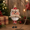 A Santa Claus hanging ornament with a rustic, weathered finish, displayed on a wooden surface with a blurred Christmas tree and holiday decorations in the background.