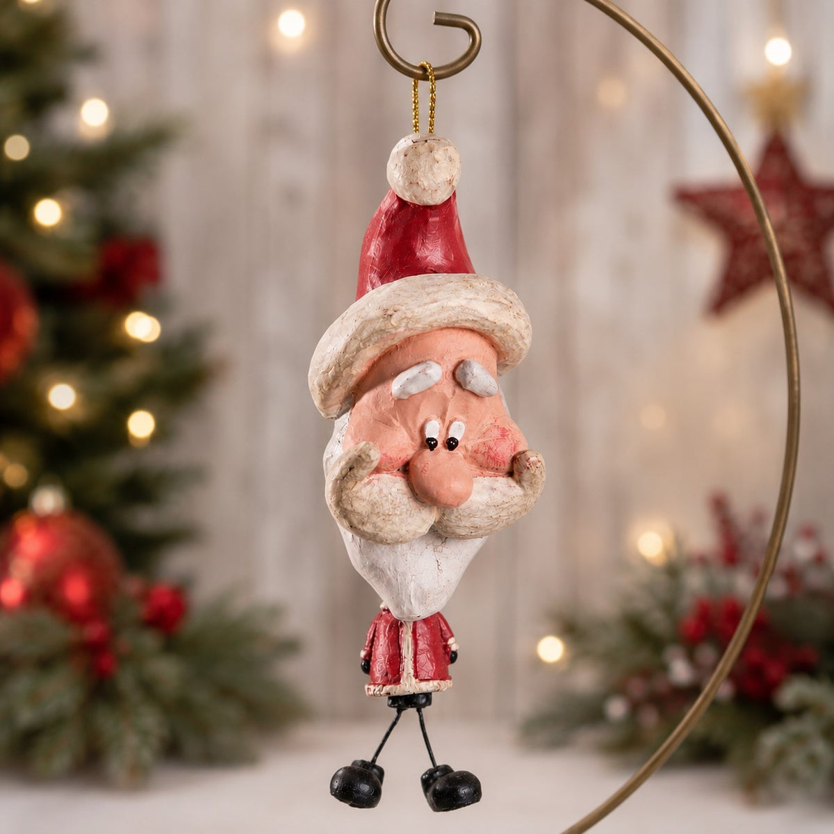 A whimsical Santa Claus hanging ornament with a elongated face, wearing a red and white Santa suit and hat, suspended by a gold  hanger against a softly blurred Christmas tree background.