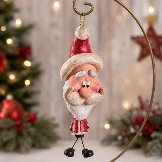 A whimsical Santa Claus hanging ornament with a elongated face, wearing a red and white Santa suit and hat, suspended by a gold  hanger against a softly blurred Christmas tree background.