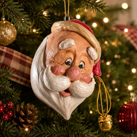A hand-painted Christmas ornament shaped like Santa's face hanging on a decorated Christmas tree. The ornament features a white beard, pink cheeks and nose, blue eyes, and a red and gold hat, with a small gold bell dangling from the side.