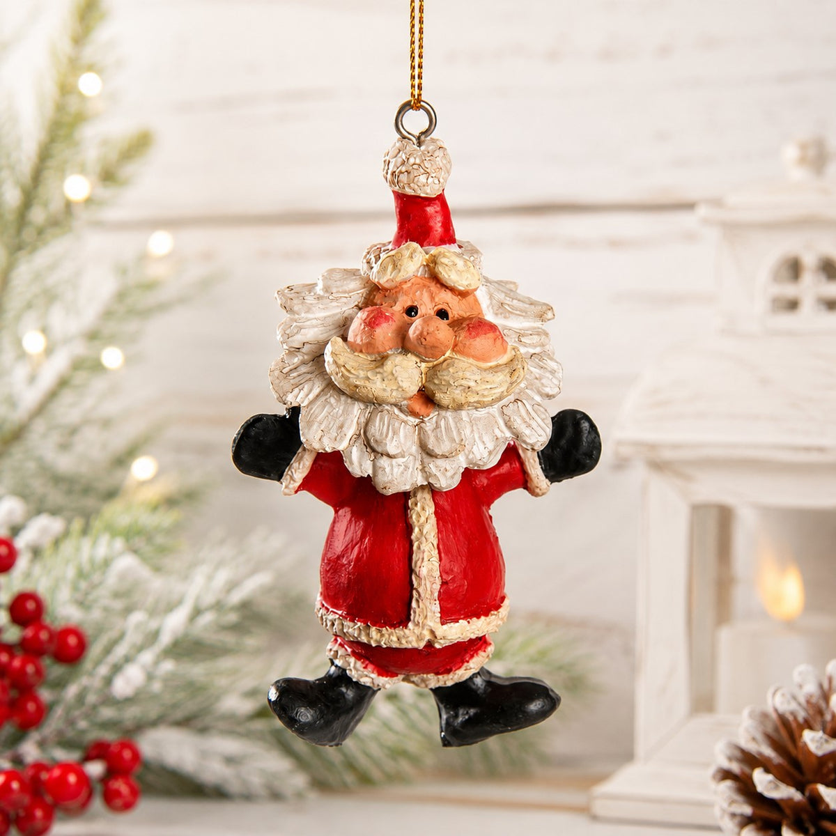 A festive Santa Claus bobble head hanging ornament with a red suit, white beard, and black boots, suspended by a gold string against a softly blurred holiday background with lights and greenery.