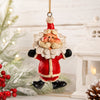 A festive Santa Claus bobble head hanging ornament with a red suit, white beard, and black boots, suspended by a gold string against a softly blurred holiday background with lights and greenery.
