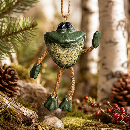 A small frog-shaped hanging ornament with a textured green and beige body, rope limbs, and black eyes, photographed outdoors among pine branches, birch trunks, moss, pinecones, and red berries.