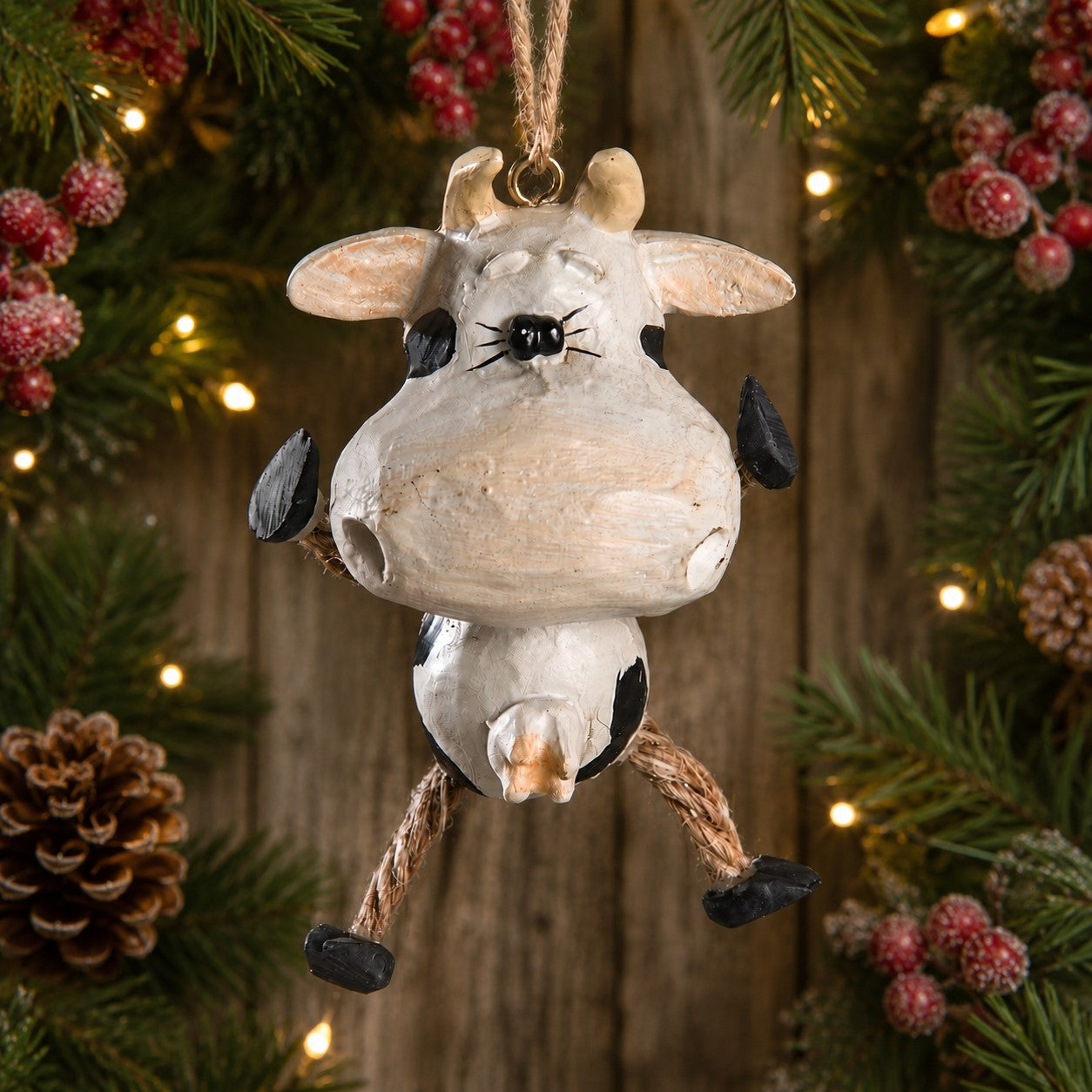 A rustic cow Christmas ornament hanging from a rope, styled against a festive holiday background of pine branches, pinecones, and warm string lights.