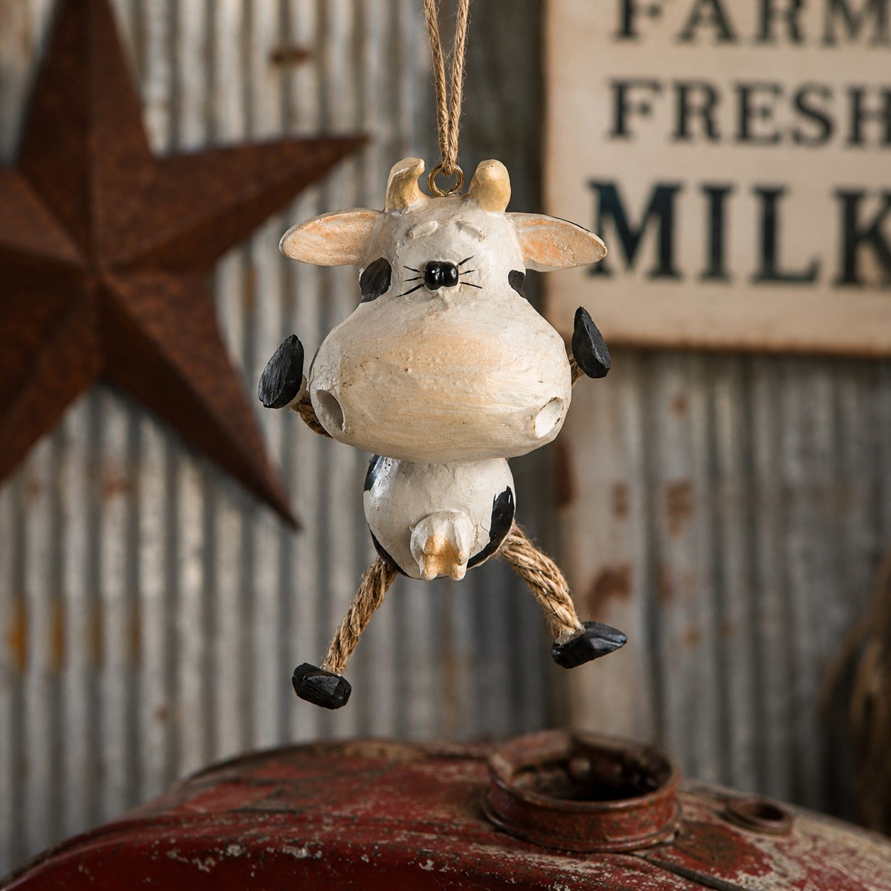A rustic hanging cow ornament with a cream-colored body, black spots, and rope legs, suspended in front of a weathered metal sign and star decoration.