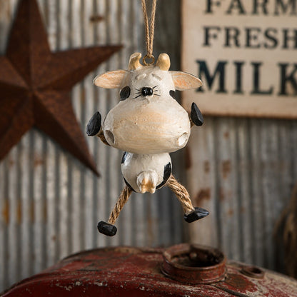 A rustic hanging cow ornament with a cream-colored body, black spots, and rope legs, suspended in front of a weathered metal sign and star decoration.