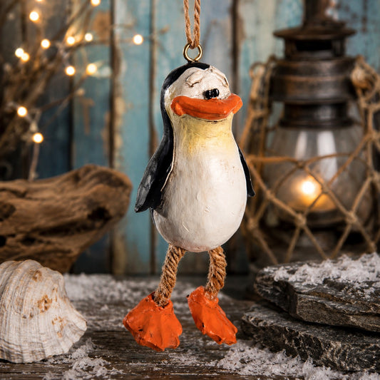 A handcrafted penguin Christmas ornament hanging from a string, featuring a distressed white and black body, orange beak and feet, and rope legs, set against a rustic nautical background with a lantern, driftwood, and seashell.