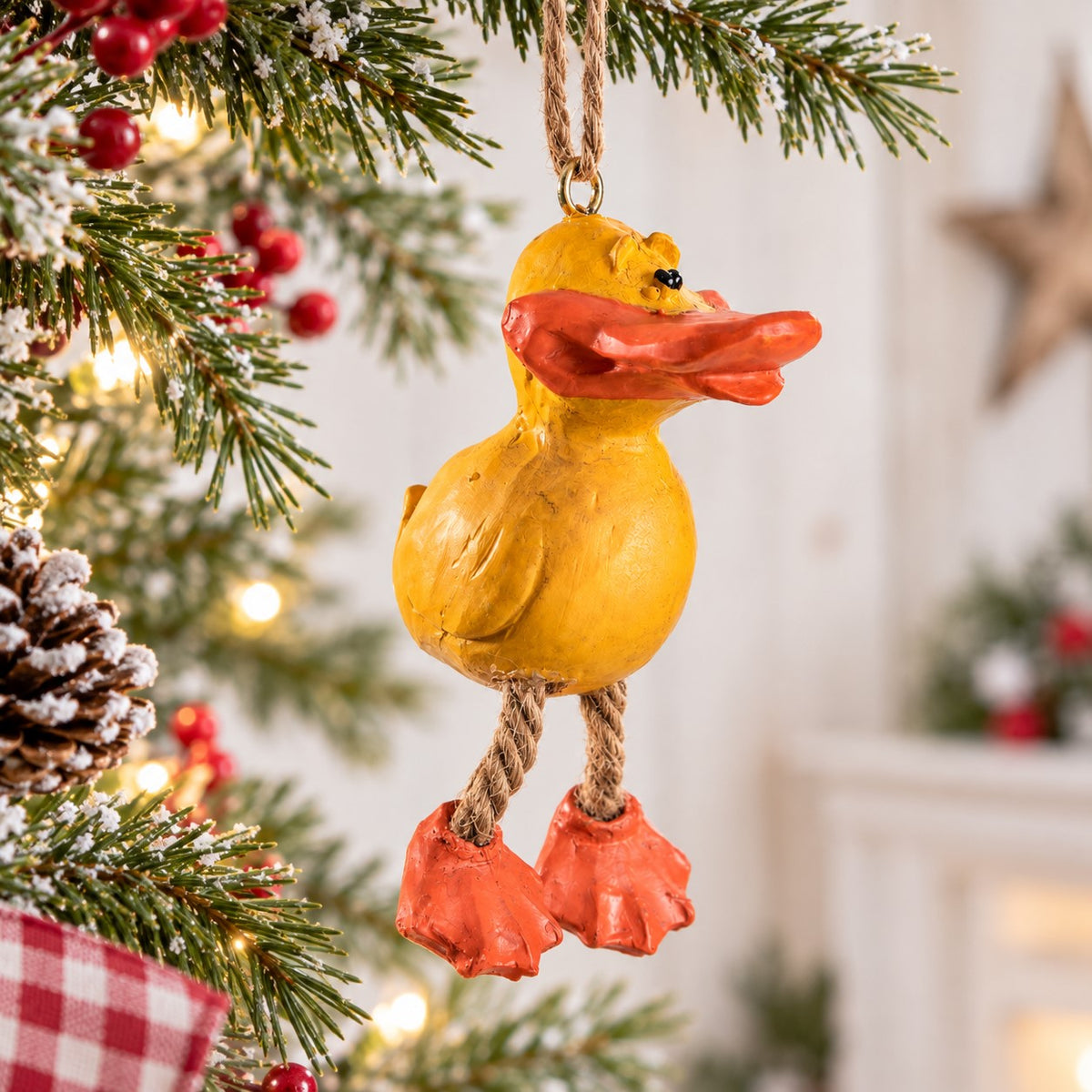 Bert Anderson yellow duck ornament with dangling orange feet hanging on a bright Christmas tree with snowy evergreen branches, red berries and warm lights.