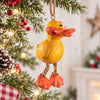 Bert Anderson yellow duck ornament with dangling orange feet hanging on a bright Christmas tree with snowy evergreen branches, red berries and warm lights.