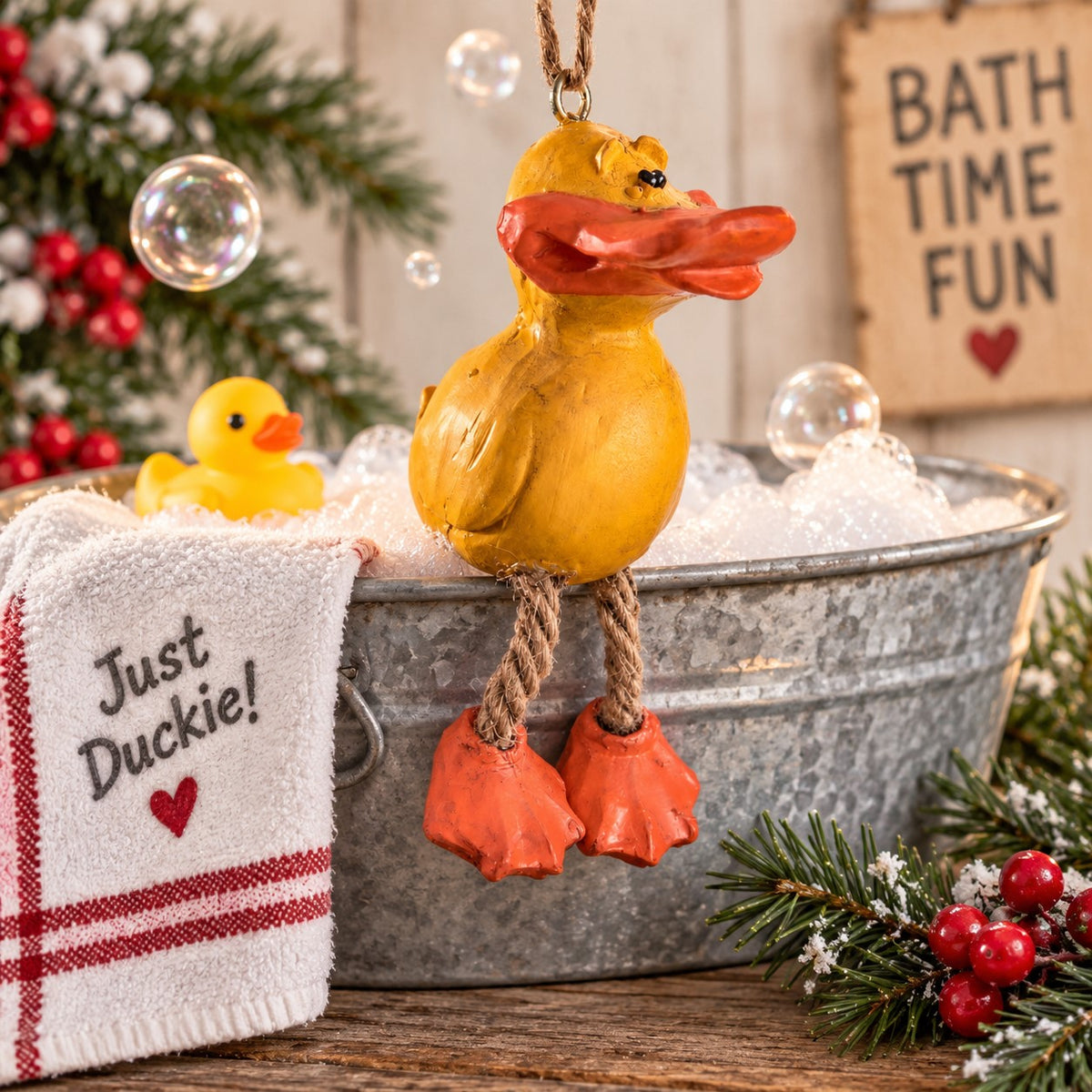 A yellow rubber duck Christmas ornament with an orange beak and feet, hanging over a galvanized metal tub filled with white foam. A small yellow rubber duck and a white towel reading 'Just Duckie!' sit beside the tub, with holiday greenery, red berries, and a wooden sign in the background.