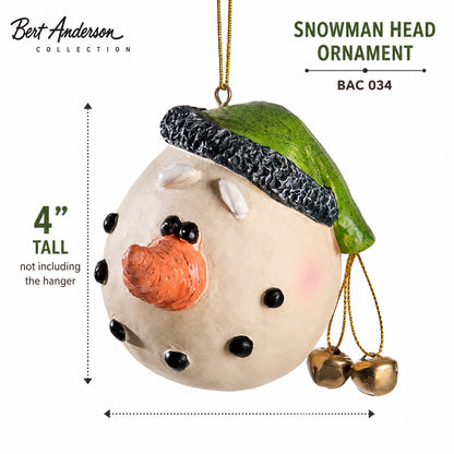 A Bert Anderson Collection snowman head ornament shown on a white background with size and product information text. The ornament is cream-colored with a green hat, black facial features, an orange nose, and small gold bells hanging from the side.