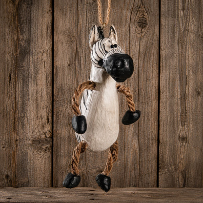 A hanging zebra plush toy or decoration made from a white stuffed body with black painted stripes, a black snout and hooves, and rope legs, shown against a rustic wooden plank background.
