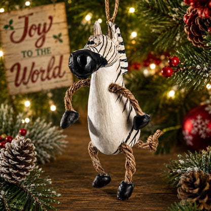 A zebra-shaped Christmas ornament hanging from a tree, styled with pine branches, pinecones, red berries, and a wooden sign that reads 'Joy to the World' in a festive holiday setting.