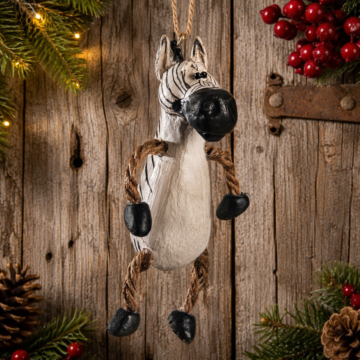 A rustic zebra hanging ornament made from a painted zebra body with rope legs and black stone hooves, displayed against a weathered wood background with holiday greenery and red berries.