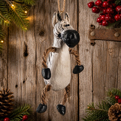 A rustic zebra hanging ornament made from a painted zebra body with rope legs and black stone hooves, displayed against a weathered wood background with holiday greenery and red berries.