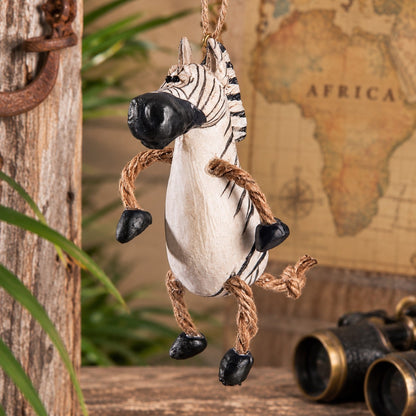 A hanging zebra decorative ornament with rope legs and black hooves, displayed on a rustic wooden surface with a vintage map and binoculars in the background.