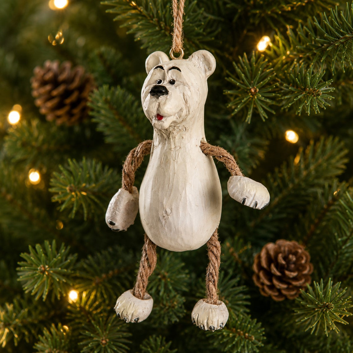 A polar bear Christmas ornament with rope arms and legs, hanging on a decorated Christmas tree with warm lights and pinecones.