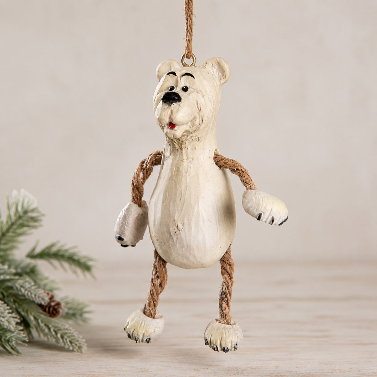 A whimsical hanging polar bear ornamnet made from resin with rope arms and legs, hanging against a neutral beige background with a pine branch in the corner.  Hanging Christmas ornament  by Bert Anderson.
