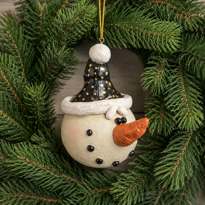 A hand-painted resin snowman ornament hanging from a gold string, nestled inside a green pine wreath. The ornament features an off-white body, a black hat with gold polka dots.