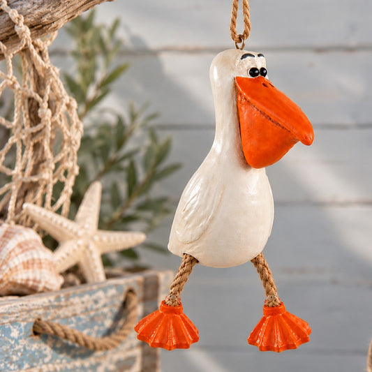 A decorative hanging pelican ornament with a  white body, bright orange beak and feet, and rope legs, shown against a softly blurred nautical-themed background with netting, shells, and a starfish.
